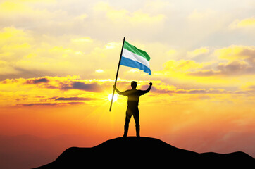 Sierra Leone flag being waved by a man celebrating success at the top of a mountain against sunset or sunrise. Sierra Leone flag for Independence Day.