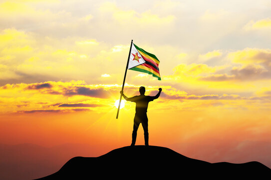 Zimbabwe flag being waved by a man celebrating success at the top of a mountain against sunset or sunrise. Zimbabwe flag for Independence Day.