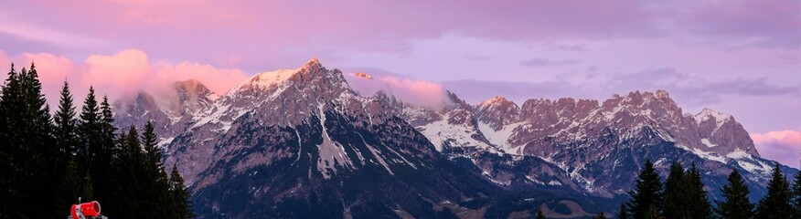 Panoramic shot of a mountain range covered with snow with pine forests in valleys at sunrise - obrazy, fototapety, plakaty