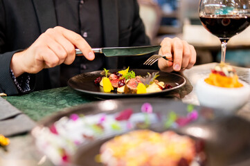 Person in black attire about to enjoy a colorful gourmet meal in an elegant setting, with a glass of red wine and additional dishes blurred in the foreground