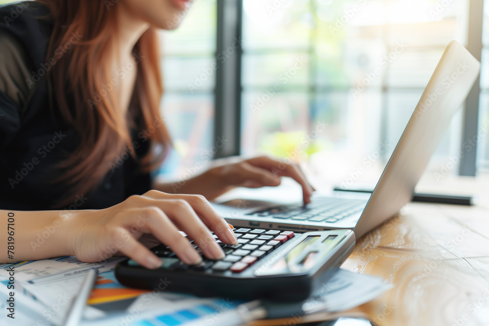 Poster a woman is using a calculator on a table with a laptop and some papers. she is focused on her work a