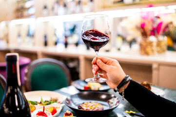 Hand swirling a glass of red wine, with a meal and wine bottle on a table in a well-lit restaurant setting