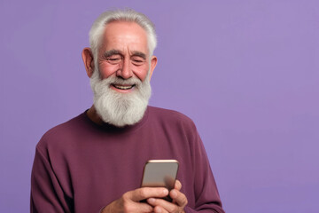 Smiling happy senior man with gray beard isolated on purple empty background uses smartphone, straight to camera
