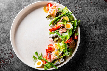Top view of a fresh salad with green leafy vegetables, sliced boiled eggs, cherry tomatoes, and pieces of fish on a white plate, placed on a dark textured surface