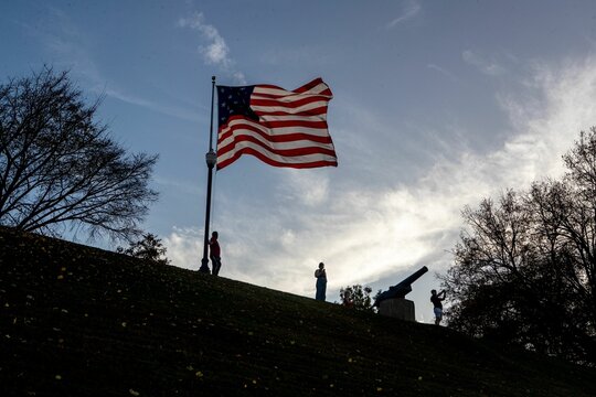 Low angle closeup of the flag of the United States of America on top of a hill with people around it
