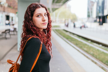 Traveler girl at public transport station waiting for the next arrival, looks at camera. She has...
