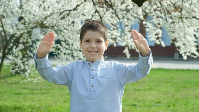 Six-year-old boy waving his hands hello or bye while standing near cherry blossom in park in spring