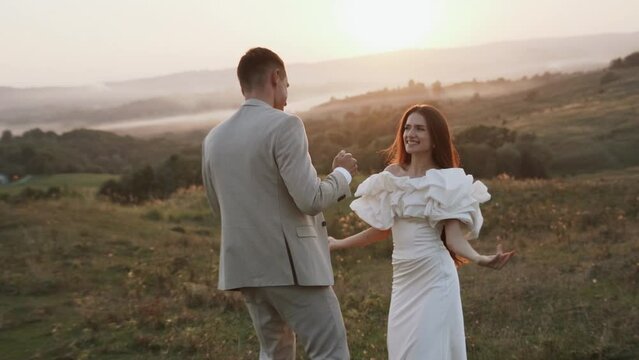 The bride and groom walk at sunset through beautiful fields and hills, laugh and dance. Wedding day concept