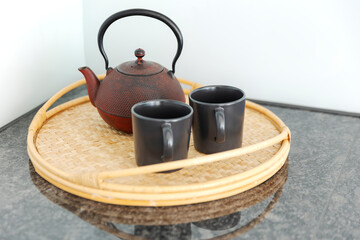 Detail of a red Chinese teapot and two black cups resting on a wicker tray, in a bright kitchen.