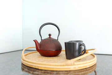 Detail of a red Chinese teapot and two black cups resting on a wicker tray, in a bright kitchen.