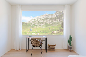 Front view of a small study with a small desk and an armchair. There is a large window overlooking a mountainous landscape with a meadow and grazing cows. - 782011365