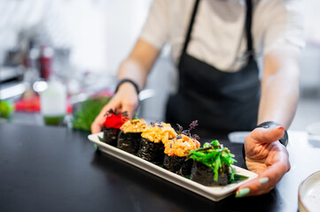 Professional chef presenting a plate of assorted sushi rolls, garnished with herbs, in a well-lit kitchen setting