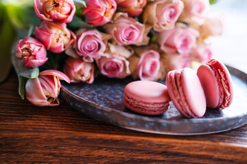 Beautiful pink roses and tulips with sweet delicacies. Sweet macaron pastries on a wooden table....