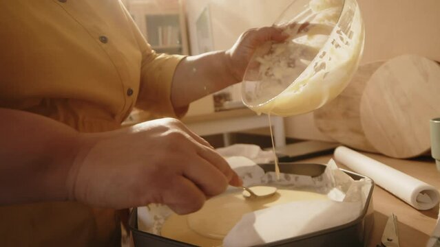 Cropped shot of unrecognizable woman pouring batter on parchment into metal cake form and spreading it over with spoon while baking homemade pastry in kitchen