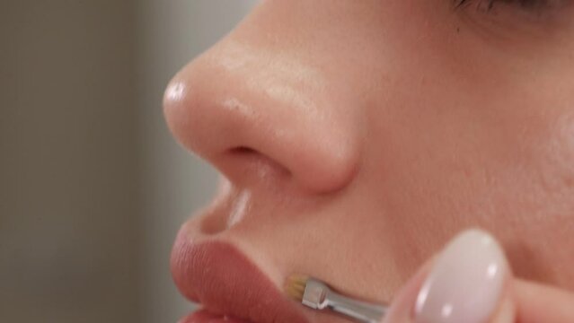 A close-up of a cosmetologist applying lip gloss with a makeup brush before permanent makeup. The cosmetologist is preparing for the permanent makeup procedure.