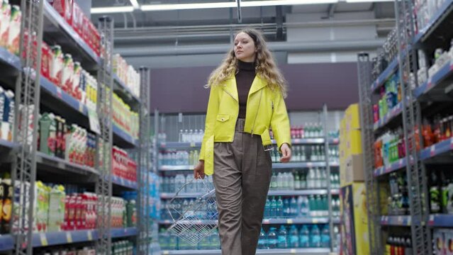 Young woman in a striking yellow jacket walks confidently down a supermarket aisle, surrounded by an array of beverages. Buying groceries at a hypermarket.