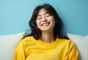 Portrait of a happy, beautiful Asian woman wearing a yellow sweatshirt lying on a sofa and smiling with her eyes closed against a blue background,
