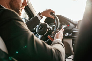 Surfing the internet by using smartphone. Man in black suit is sitting in the car, driving