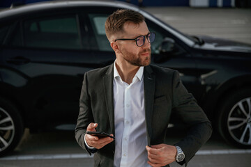 Smartphone in hand. Businessman in suit is near his black car outdoors