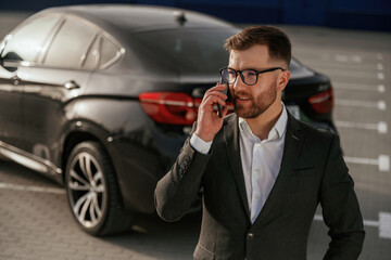 Talking by using phone. Businessman in suit is near his black car outdoors