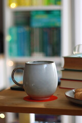 Cup of tea or coffee, pile of books, plate of cookies, reading glasses, e-reader and pen on the table. Colorful rainbow bookshelf in the background. Selective focus.