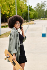 A young African American woman with curly hair multitasks by holding a skateboard and talking on a cell phone in a skate park.