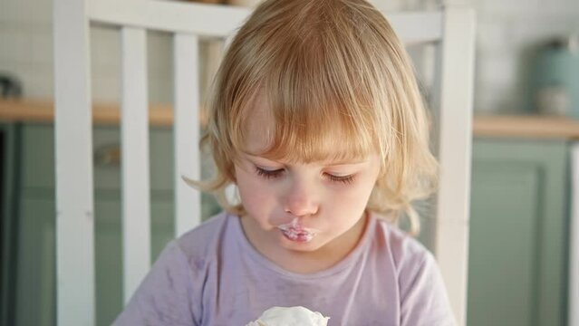 Baby girl enjoying ice cream. Pretty little toddler eating an ice-cream indoors, at home. Dining room background. Small child eats plombir and cream messy on her mouth. Cute kid with tasty sweet food.