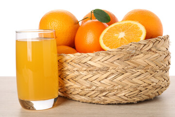 Fresh oranges in wicker basket and glass of juice on light wooden table against white background