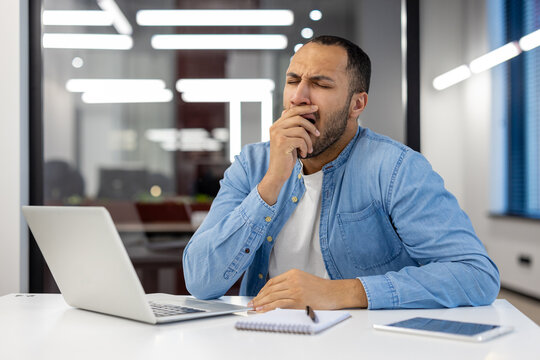 A tired young Muslim man in a blue shirt sits at a desk in a modern office and yawns while covering his mouth with his hand