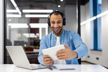 Smiling young Latin American man sitting at a desk in the office wearing headphones and...