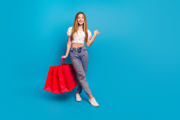 Full length portrait of lovely girl hold bags direct finger empty space wear top isolated on blue color background
