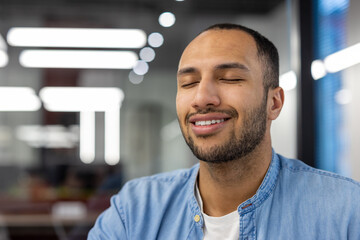 Close-up photo of a calm and smiling young Arab man who is in the working office and rests and...