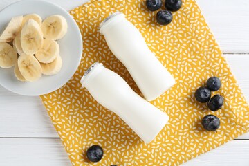 Tasty yogurt in bottles, banana and blueberries on white wooden table, top view