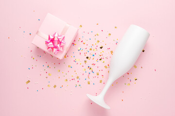 White champagne glass and gift box with confetti on pink background. Minimal party concept.