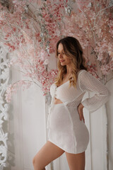 Fototapeta premium Beautiful pregnant woman in special white beach dress. Advertisement of comfortable pregnancy Happy blond girl stand in room with flowers. Caucausian woman portrait