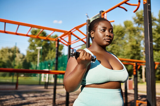 Curvy African American woman in blue top lifting a dumbbell outdoors, embodying strength and confidence.