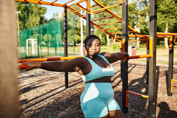 An African American woman in sportswear strikes a pose in front of a playground, showcasing body positivity.