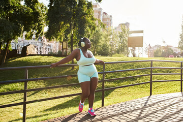 A curvy African American woman in headphones is leaning on a fence, wearing a white top and blue shorts, lost in the music.