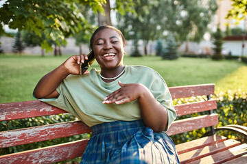 Plus size African American woman in casual attire, sitting on a bench outdoors in the summer, talking on a cell phone.