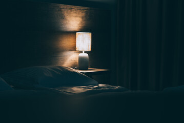 The cozy bed in the hotel at night with yellow night lamp. The light from the lamp reflects on the wooden wall. Toned image with copy space for you text.