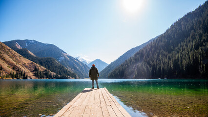 A man stands on a wooden pier looking out at a tranquil mountain lake, surrounded by forested...