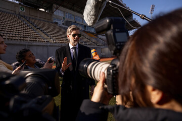 Interview of football team director at press conference for TV news. Organization representative answering questions and giving interview outside a soccer stadium. Journalists crowd on press campaign.