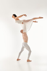 A shirtless young man and a woman in a white dress gracefully dance together, incorporating acrobatic elements, against a white studio backdrop.