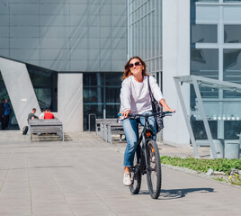 A happy smiling caucasian woman in fancy sunglasses riding a bicycle on the city modern street. Ecology sustainable transport and happy people concept image.