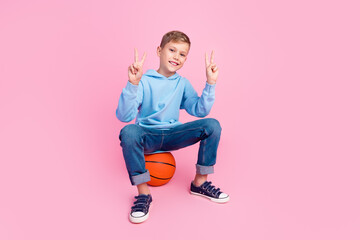 Full size photo of little cheerful boy sit basketball show v-sign empty space isolated on pink color background