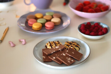 Plate of pastel macarons, cookies and chocolate, cup of tea of coffee, glass of bubble water, various berries, books and accessories on the table. Selective focus, pastel colors.
