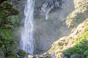 Sunlit waterfall cascades through lush NZ foliage, a serene Arthur's Pass gem