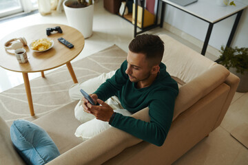 Young man sitting on couch and texting friends - 782090167