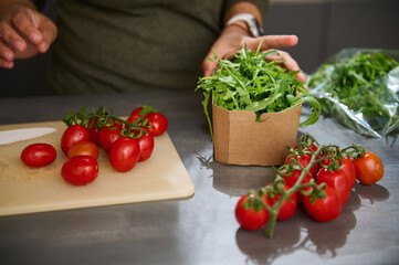 Woman stand at kitchen counter with fresh ingredients in eco friendly recyclable cardboard bags. A bunch of red ripe tomato cherry and arugula leaves for healthy salad. Diet. Vegetarianism. Vegan food