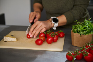 Close-up woman hands chopping cherry tomato on a cutting board, preparing healthy salad with fresh arugula leaves, standing at kitchen counter. Culinary, diet, healthy eating and food concept